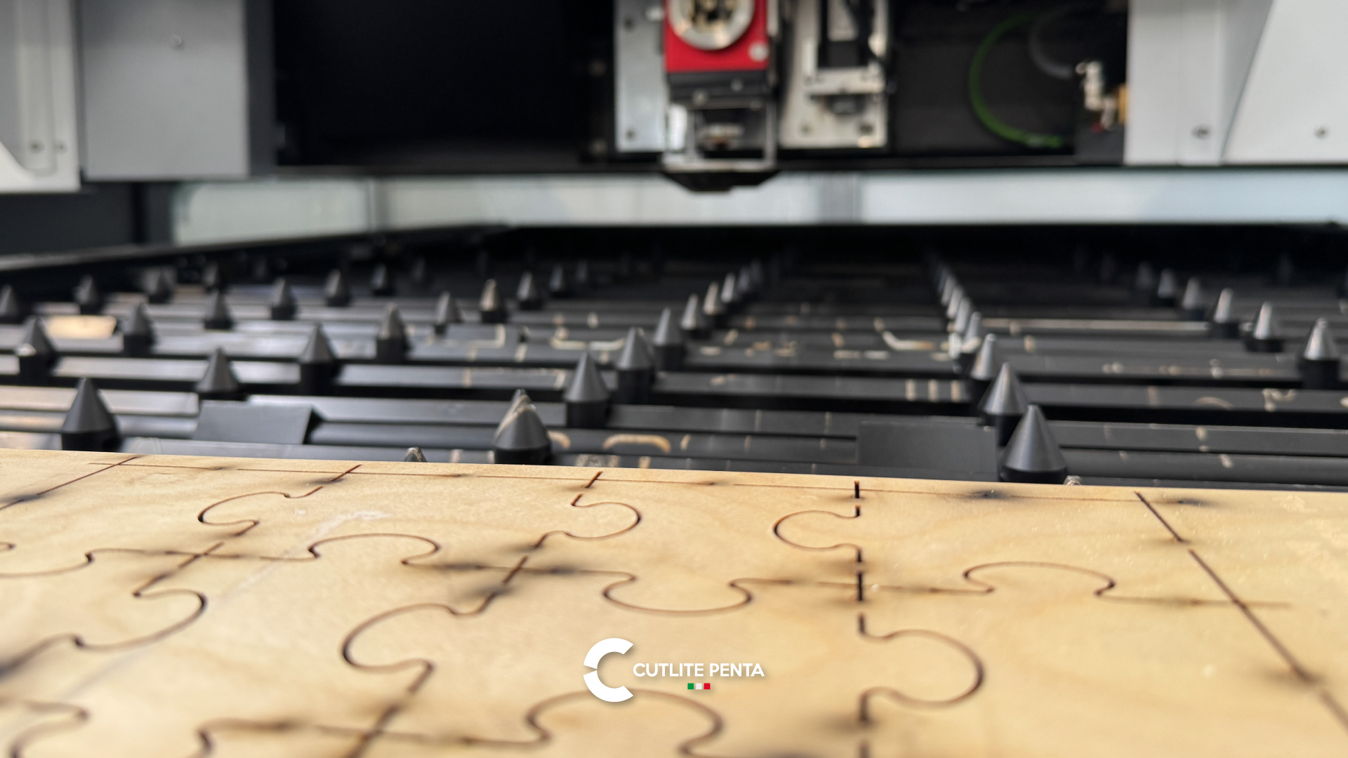 Una tavola di legno sul banco della macchina laser, con le fessure incise dal laser che disegnano un motivo a puzzle; sopra, la testa di taglio