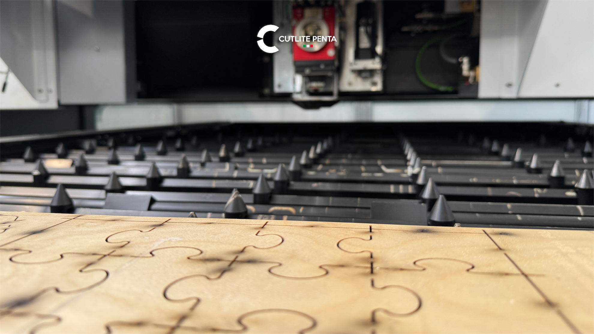 Un tablero de madera en la mesa de la máquina láser, con las ranuras grabadas por el láser formando un motivo de puzle; arriba, el cabezal de corte