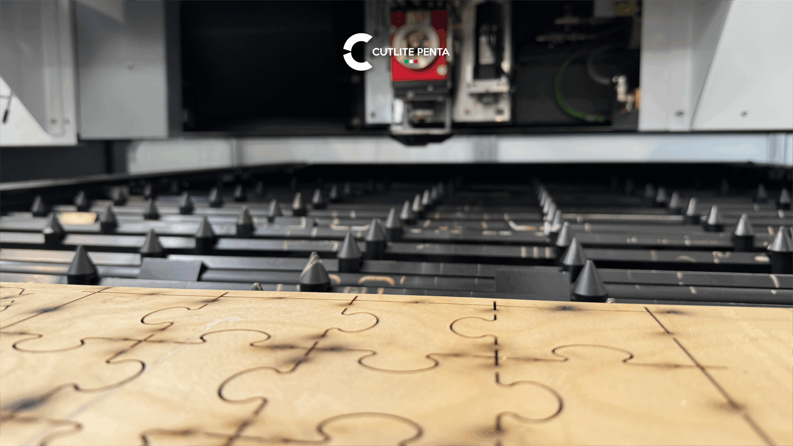 A plywood board on the laser machine bed, with laser-cut slots forming a jigsaw pattern; the cutting head above