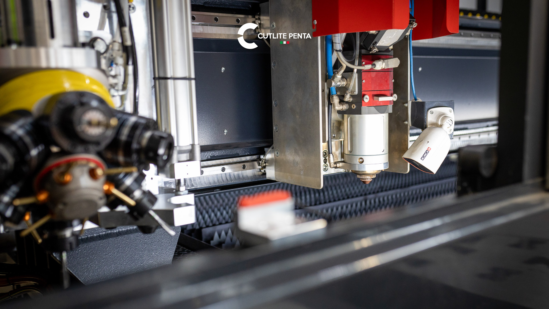 Cabeçote de corte com carcaça vermelha e bico de cobre no interior da cabine de proteção de uma máquina laser Cutlite Penta, com uma câmera voltada para a área de trabalho, grade de suporte e ferramentas em estação em primeiro plano