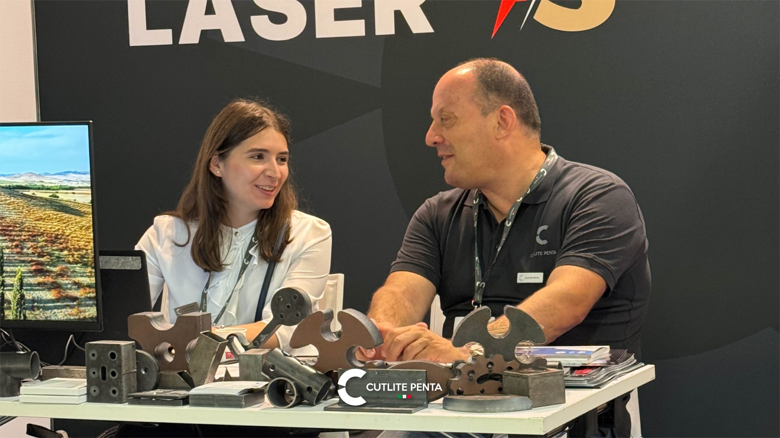 Lucio Verdi talking with a visitor at the table of cut samples, in front of the LASER VS PLASMA graphic