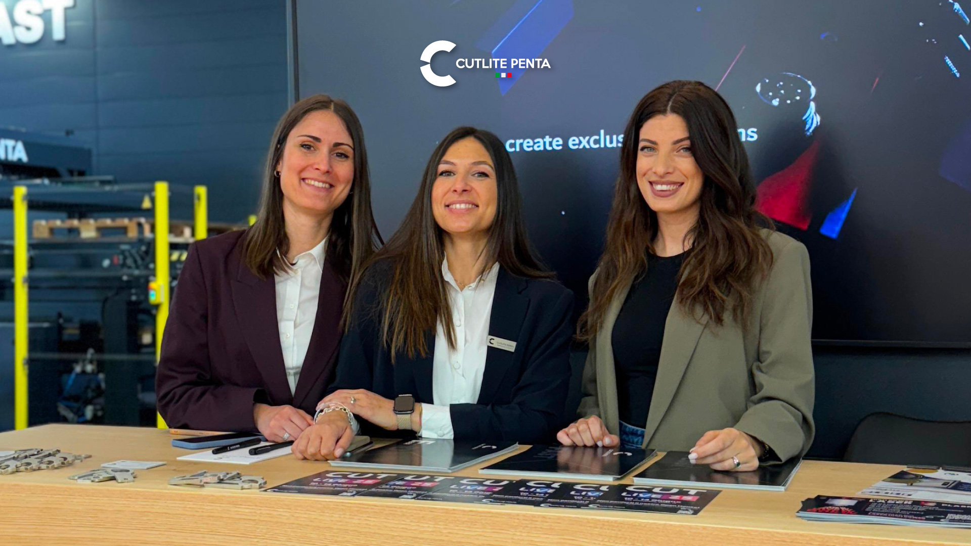Carolina Delibassis, Alessia Rugi et Elvira D'Afiero du service commercial de Cutlite Penta au comptoir d'accueil pendant un événement dans l'entreprise, avec des échantillons de découpe et des flyers sur la table